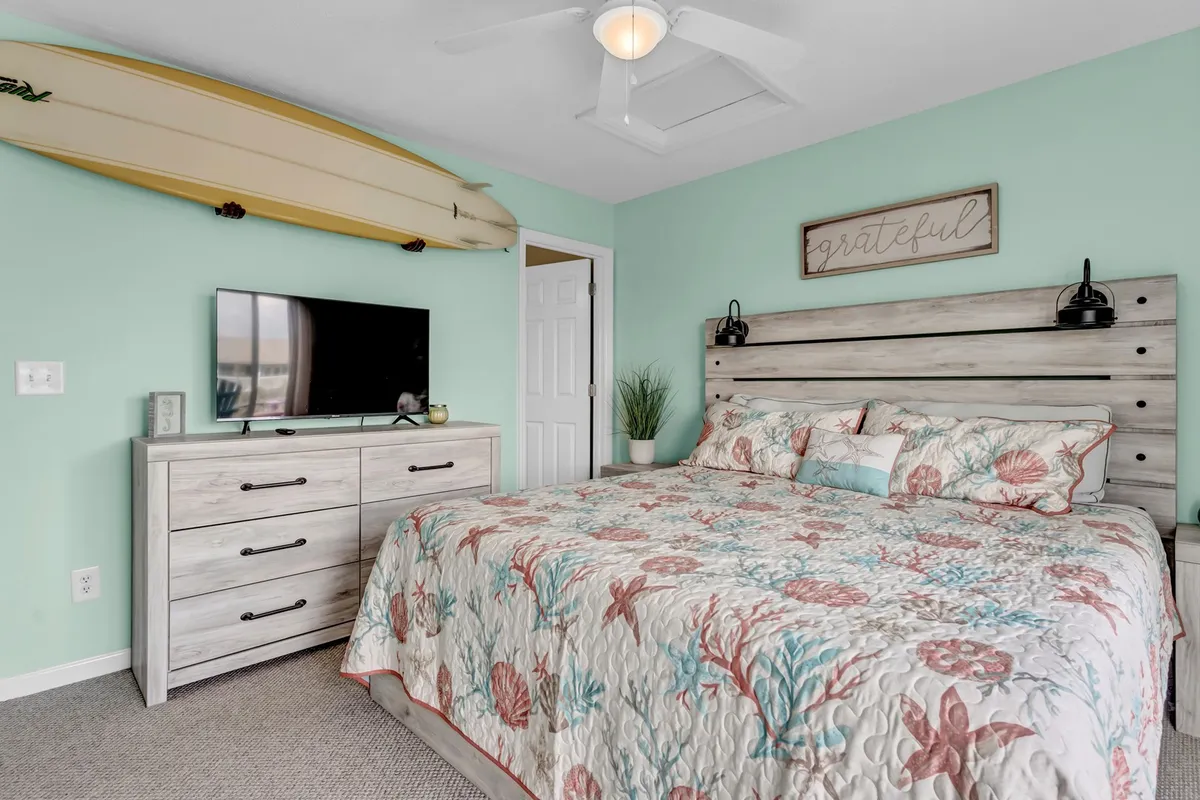 A casually inviting bedroom with a surfboard accent on the wall, a well-made bed, dresser, and wall-mounted flat-screen TV. Large windows fill the space with light, suggesting a comfortable setting for relaxing or enjoying a movie night after a day at the beach.