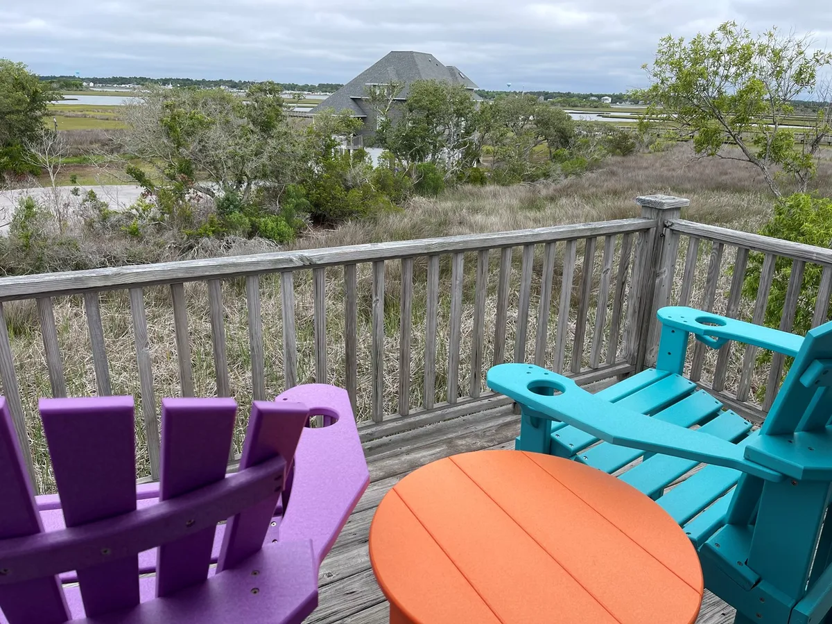 The image showcases a cozy deck with colorful Adirondack chairs (purple and turquoise) and an orange side table facing a scenic natural view, likely overlooking marshland. The setting captures a relaxing, casual vibe perfect for unwinding while enjoying the outdoors. The atmosphere is bright and inviting, suggesting a place where guests can gather for conversation, watch the sunset, or sip morning coffee.