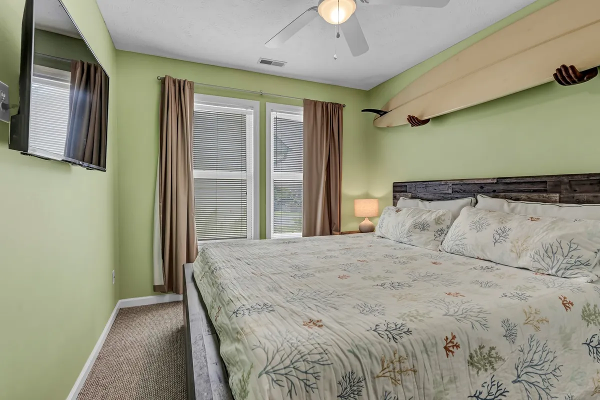 Inside, the room features a cozy bed with floral bedding, a surfboard on the wall, and large windows letting in natural light. The space feels tranquil and welcoming, ready for new guests to settle in and relax.