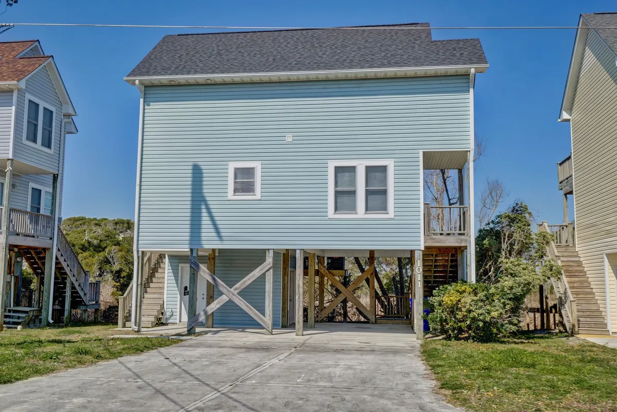 A light blue raised house nestled among lush greenery, with a welcoming porch and steps leading up from a spacious lawn. The home’s exterior radiates a relaxed coastal vibe, perfect for gatherings and outdoor relaxation. The setting suggests a peaceful, sociable environment close to local amenities.