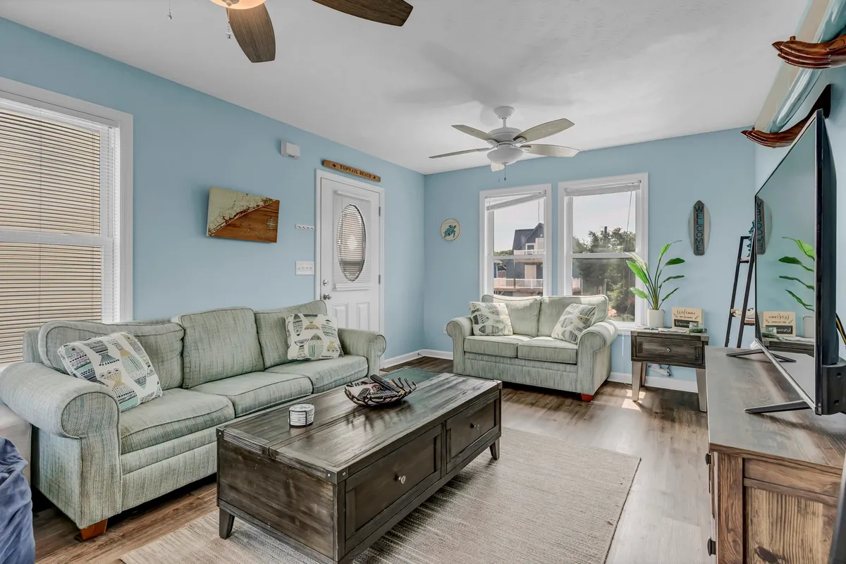 A cozy living room filled with natural light, featuring a plush sofa, coffee table, and large flat-screen TV. Generous windows illuminate the space, and the arrangement invites relaxation and group gatherings, perfect for socializing or enjoying a movie night.