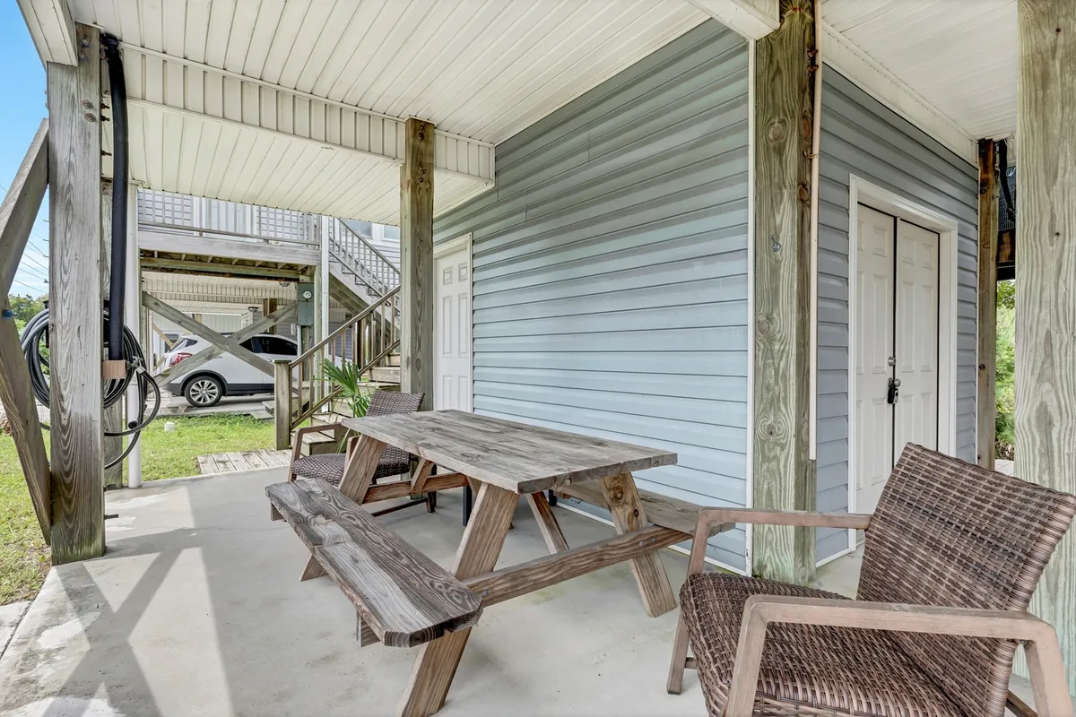This image captures an inviting outdoor area with a wooden picnic table, woven chairs, blue house siding, and stairs leading up to the home. The space is designed for outdoor dining and gatherings, reinforcing a welcoming atmosphere for new arrivals.